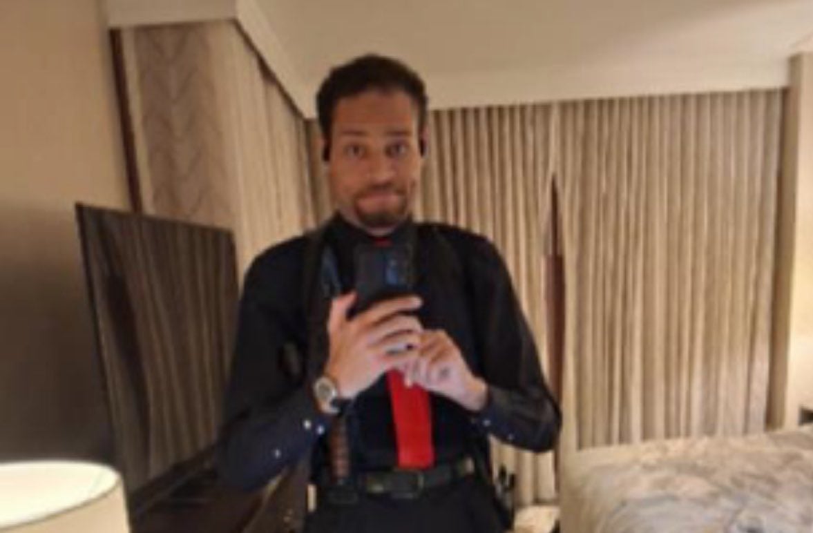 Με μαύρο πουκάμισο, κόκκινη γραβάτα και ζωσμένος με όπλα ποζάρει χαμογελώντας πριν πυροβολήσει